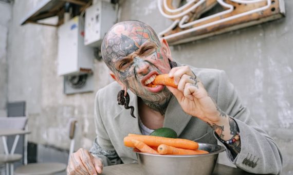 Tattooed man eating fresh carrots with a playful expression in an urban outdoor setting.