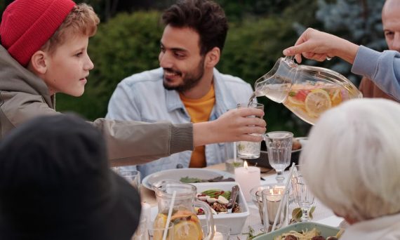 Faceless female pouring fruit beverage into glass of boy in warm cloth while cheerful multiethnic family having dinner outdoors